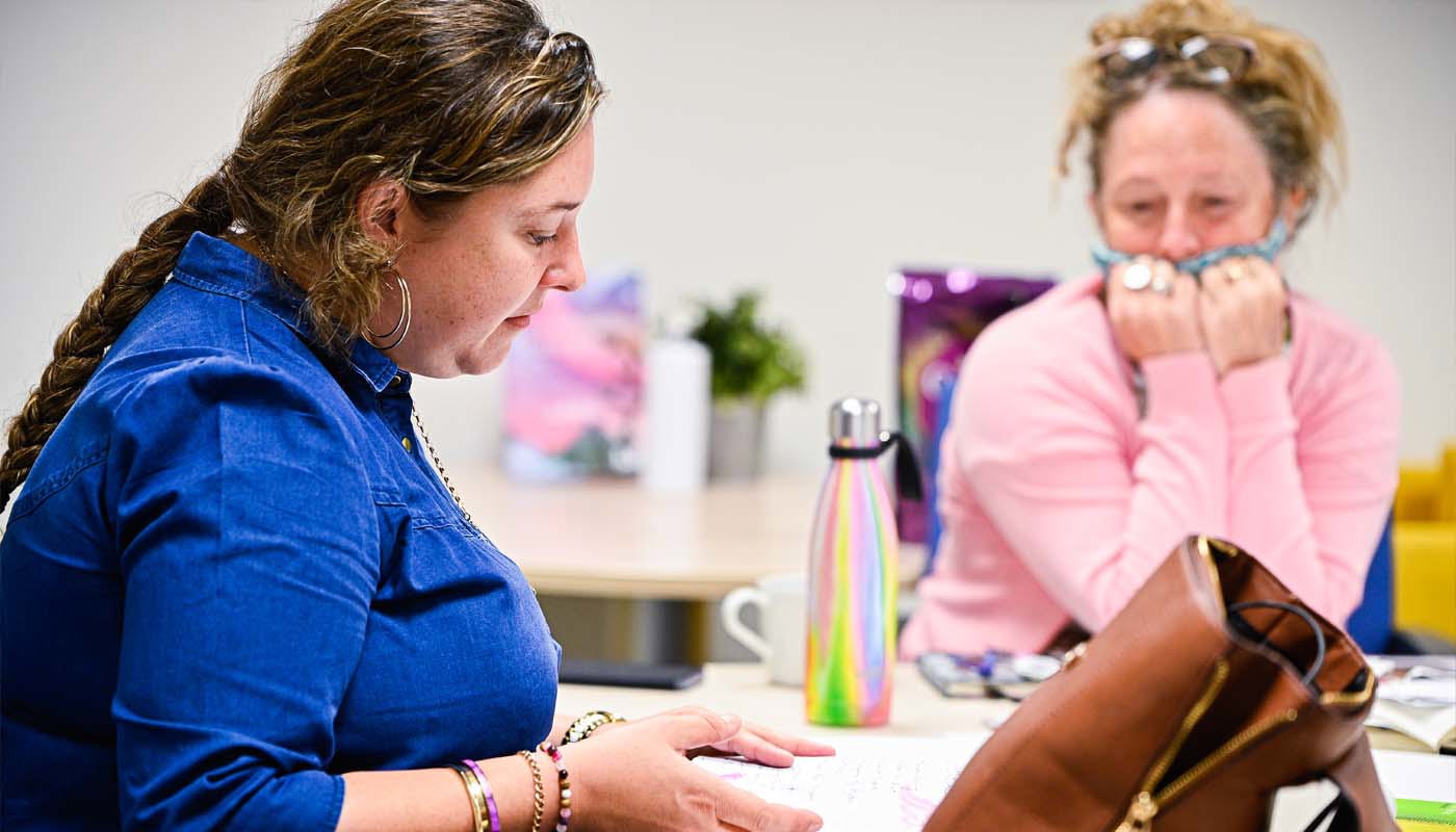 8.	Two women sit a table. One is a young woman who has a notebook on the table in front of her. She is looking down at the notebook. The other woman is middle-aged and is looking towards the notebook. 