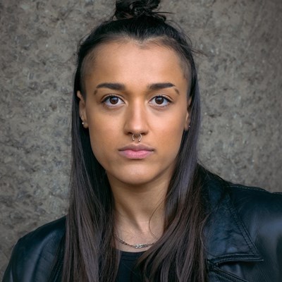 Aisha-May Hunte's headshot. Aisha is a young, mixed race person. They have dark hair, half tied up in a bun at the top of their head. They are looking directly into the camera and they are not smiling. They are wearing a black leather jacket. 