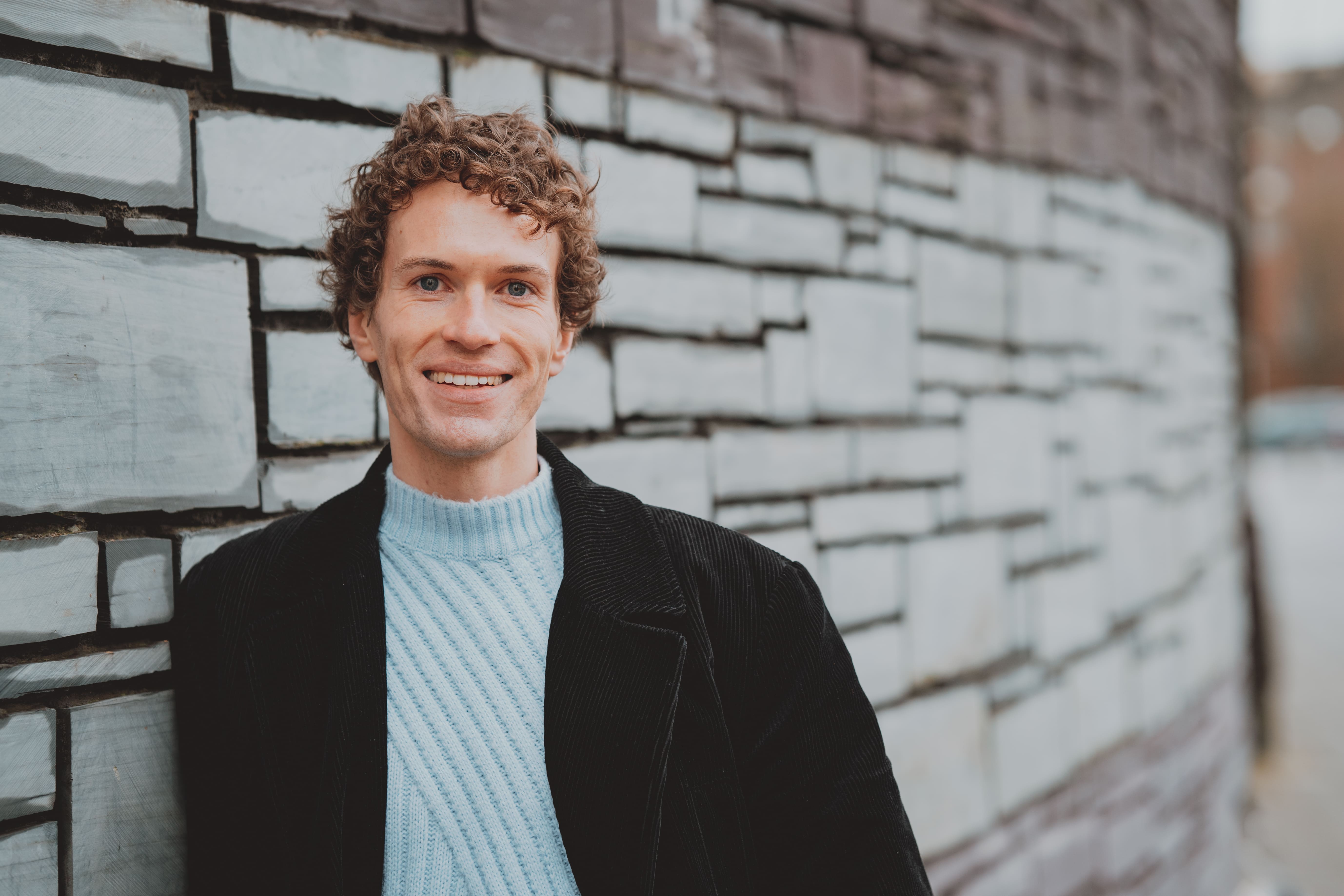 Exterior shot of a young man. He has short curly brown hair. He is wearing a light blue knitted jumper with a black blazer on top. He is leaning against a slate wall. He is staring directly at the camera and smiling with his teeth showing.