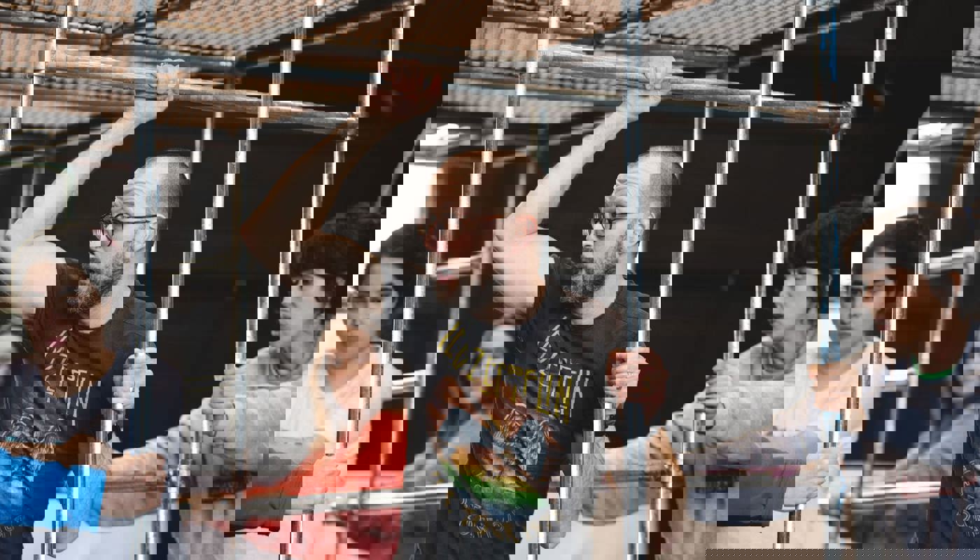 4 people stand around a metal structure built from poles. A man stands inside the structure and holds a pole above his head and another pole below this. A young woman is stood behind him with her arms stretched out. Behind them on the edges of the structure, two women are holding the poles and looking towards the other two. Nobody is smiling.