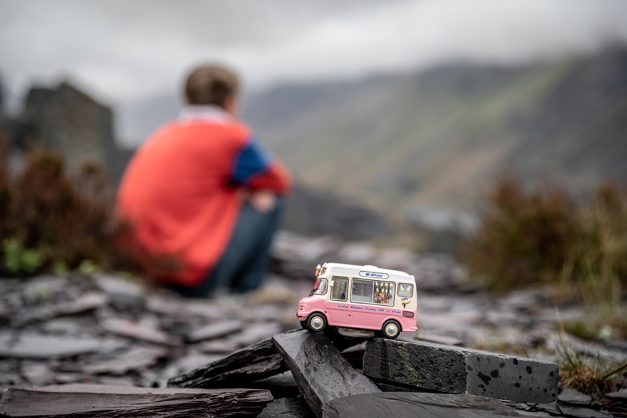 Exterior shot. The sky is grey and there are mountains in the background, unfocused. In front and in focus there is a pink and white small model ice cream van placed on a pile of small slates. Behind it out of focus a boy is sitting down with his back to the camera.