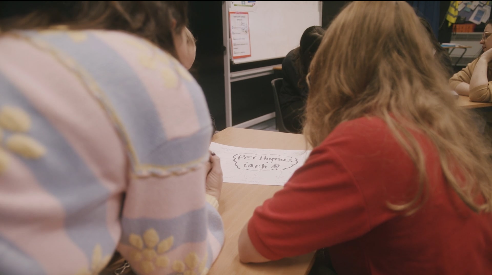 A woman in a stripey pastel cardigan leans over a table next to a young person wearing a school uniform. A piece of paper is on the table in front of them with writing which reads 'Healthy relationship'