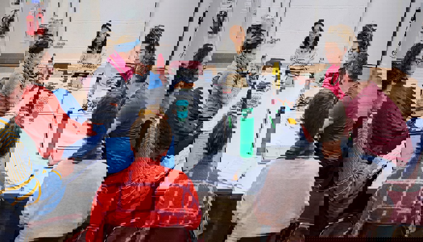 A room full of young people sat around a table. They are a mix of men and women. On the table there are scripts, water bottles, coffee cups and various other pieces of paper. Many of them are smiling and looking at one of the people who is reading something on his phone.