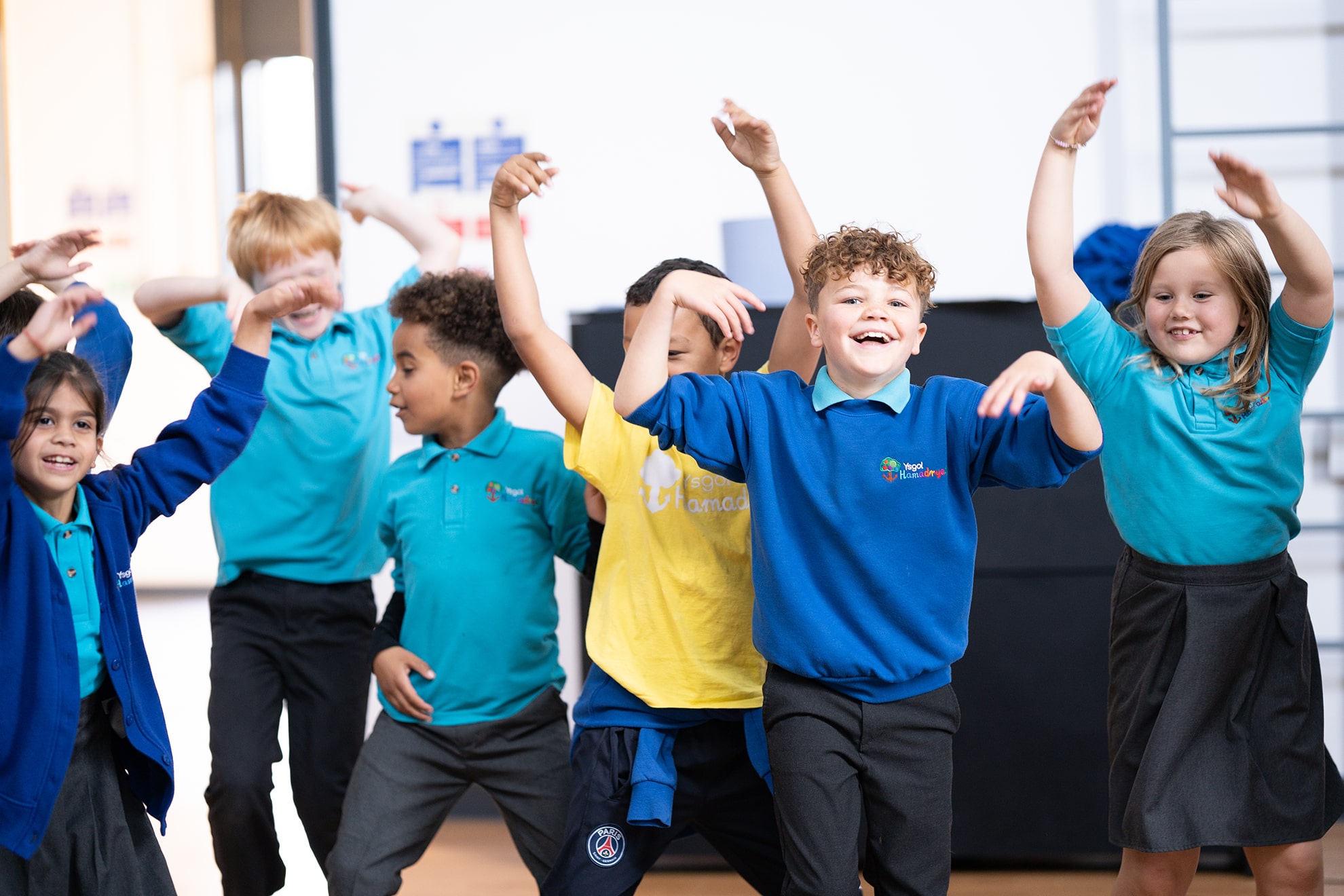 A group of young children during the Swyn workshops. They are stood together smiling, with their arms stretched up. They are all dressed in school uniform.