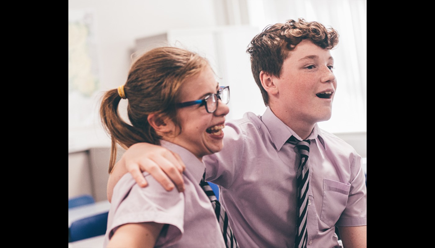 An image of two school children. One boy and one girl with the boy's arms around the girl. They are both laughing and smiling.