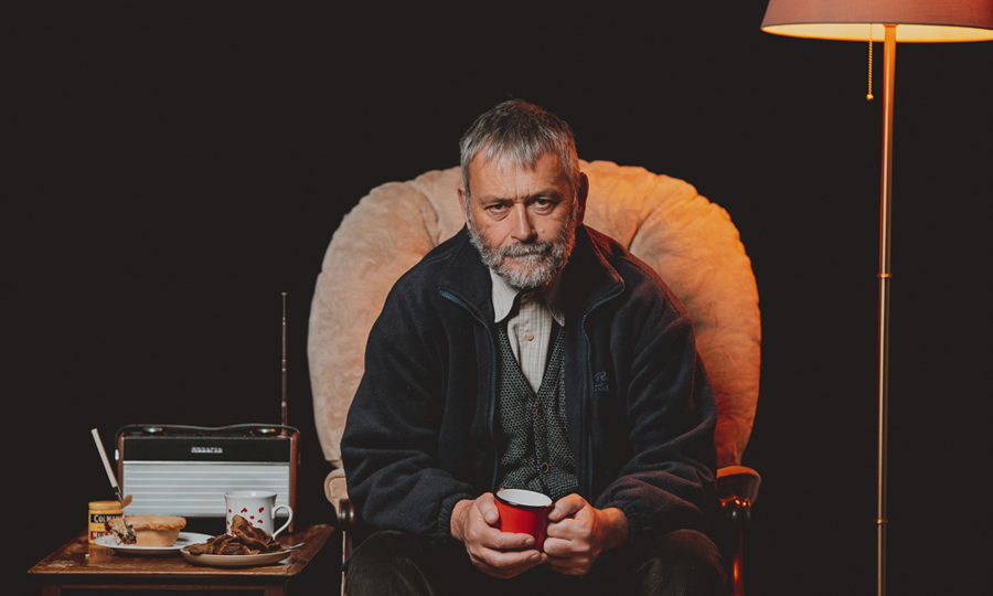 Huw Fyw poster image - a man with a grey beard sits in an armchair holding a cup of tea.