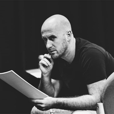 Image of Kieran Bailey. He is a white male. He is bald and has a very short stubble on this chin. He is resting his head in his hand and holding a script in his other hand