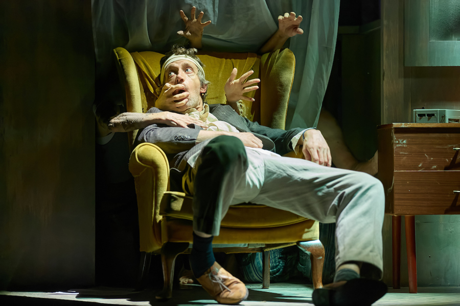 A man with a bandage on his head sits in a yellow wing armchair. There are hands reaching through the chair and around it. One hand covers his mouth,