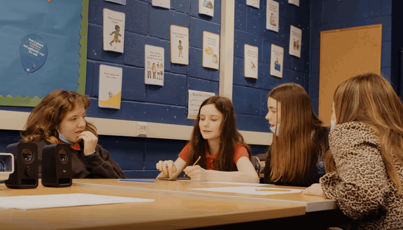 3 young girls in school uniform and a young woman in formal clothing sit around a table. 2 of them are wearing masks lowered down around their chin. Two of their mouths are open mid-conversation. One of the girls is holding a pencil, and they are looking at a piece of paper that is on the table.