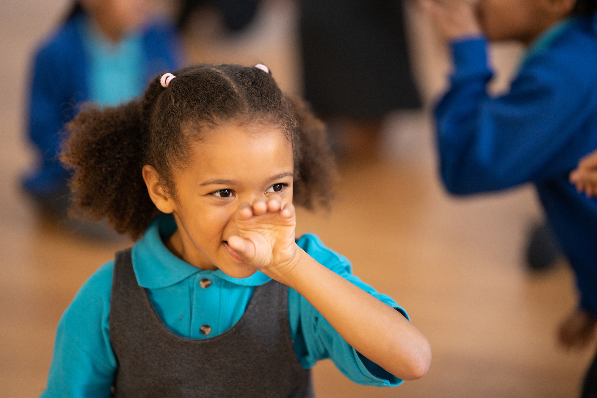 Image of a young black girl with afro hair tied  into two ponytails at the back of her head. She is wearing a light blue polo shirt under a grey pinafore. She holds her hand to her mouth her palm half closed.