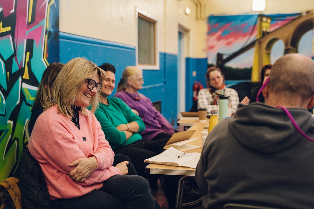 Group of people sat around a table. One woman is particularly in focus and she is staring at something in front of her and smiling. There are pieces of paper and pens on the table in front of them. On the walls around them there is various colourful pieces of graffiti.
