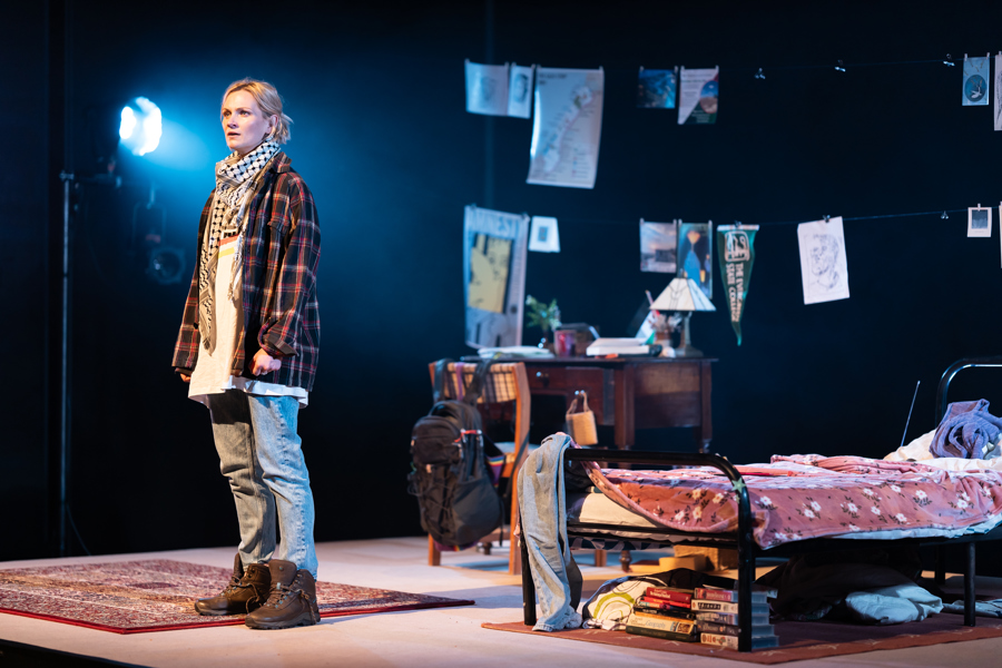 Production image for Fy Enw i yw Rachel Corrie. A woman is standing on stage wearing a check shirt, tshirt, jeans, heavy boots and a keffiyeh. Behind her is a bedroom scene.