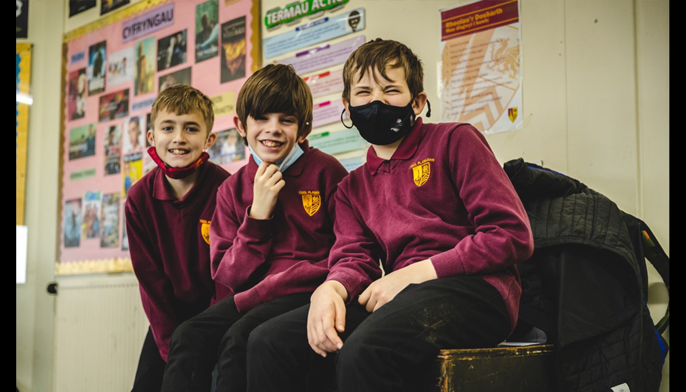 Three young male students are sat together on a table. They all wear a school uniform and one is wearing a black face mask. They are all smiling and looking directly into the camera.