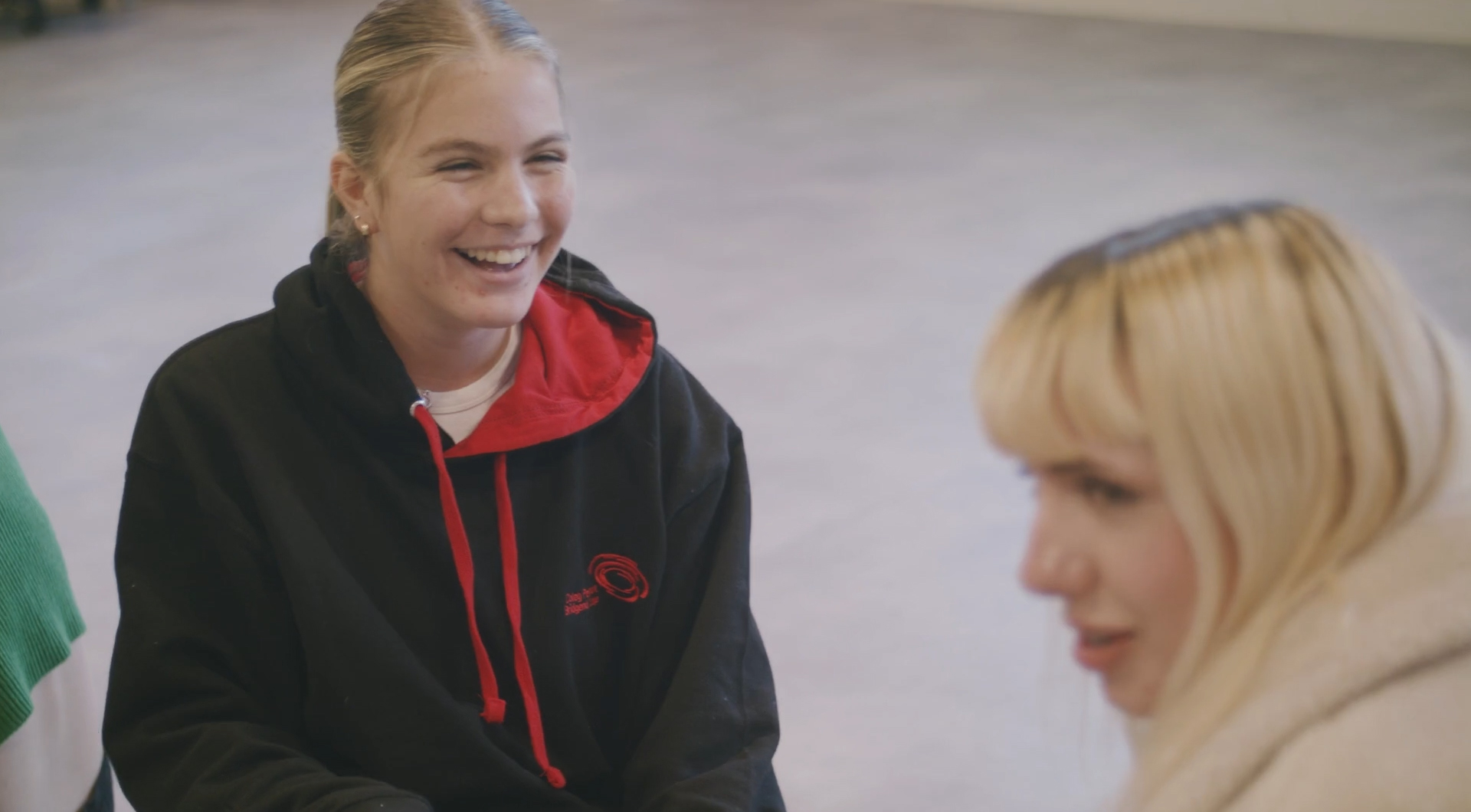 Two young white blonde women are sat on the floor having a discussion. One is smiling widely, and the other appears mid sentence.