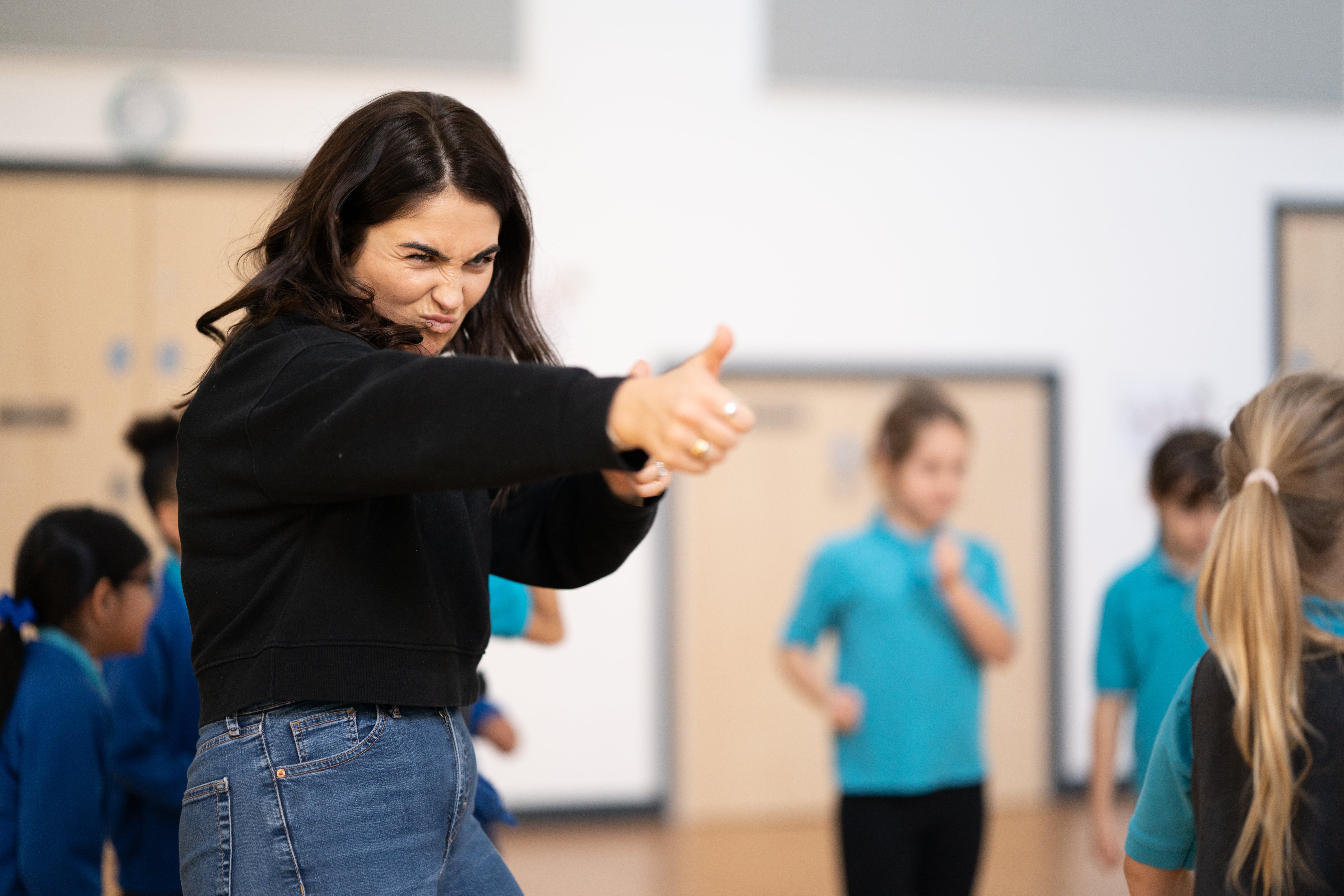 Image of Sian Elin James in workshop. Children in school uniform are stood in the background. Sian Elin is a white woman in her twenties with long dark hair. She is looking towards someone and giving them a big thumbs up.