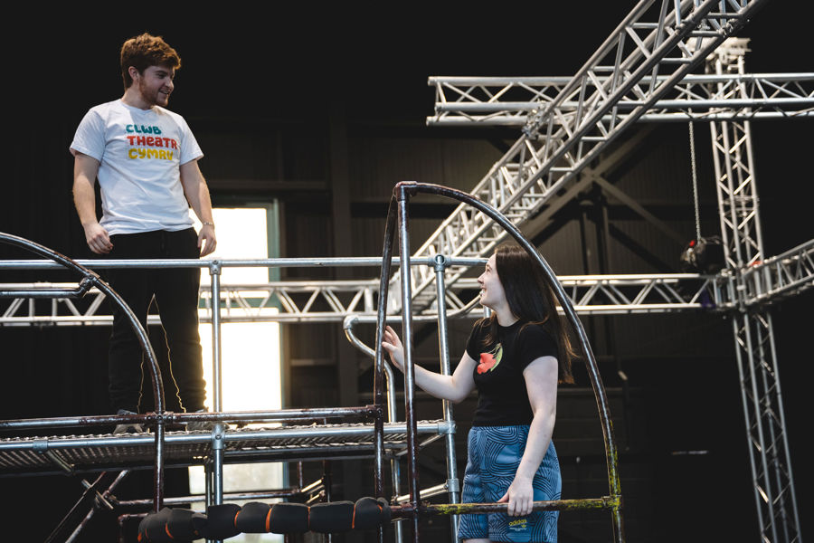 A young man stands on top of a metal structure built from poles. His mouth is slightly open and he looks down towards a young woman who is leaning on a small ladder built into the side of the structure. She is smiling and looks up at him.