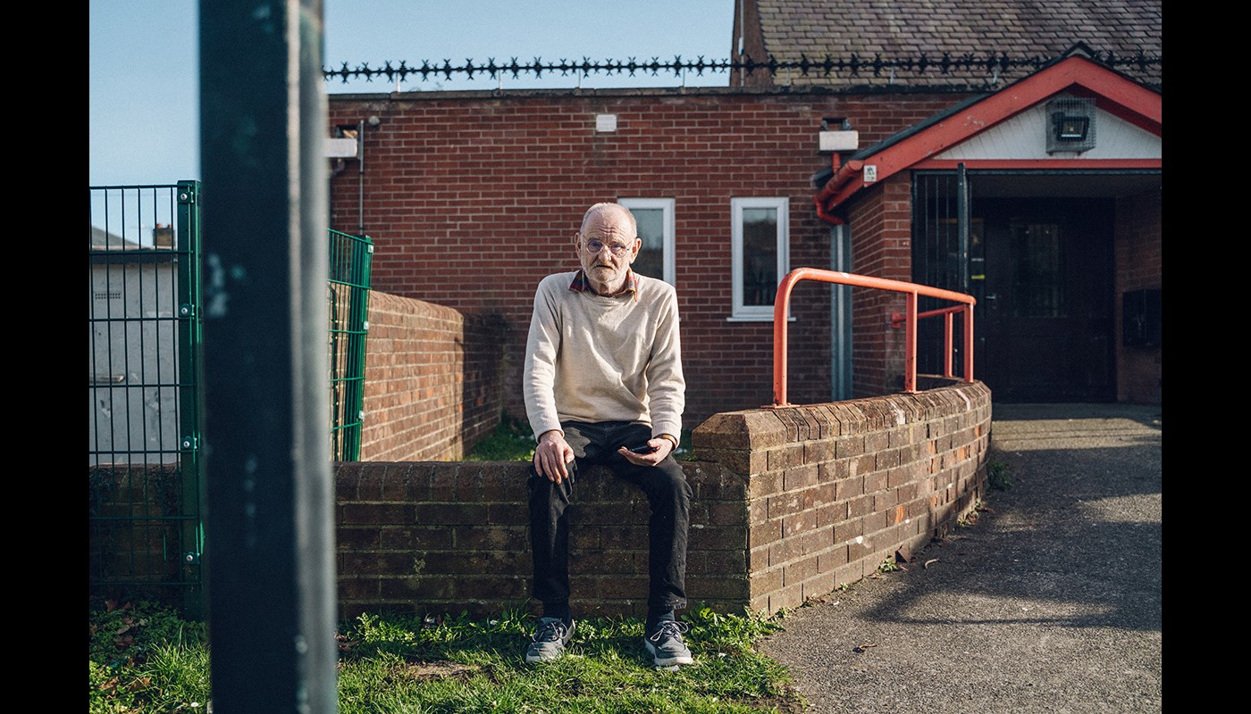 An elderly man sits on a low brick wall outside a community centre. 
