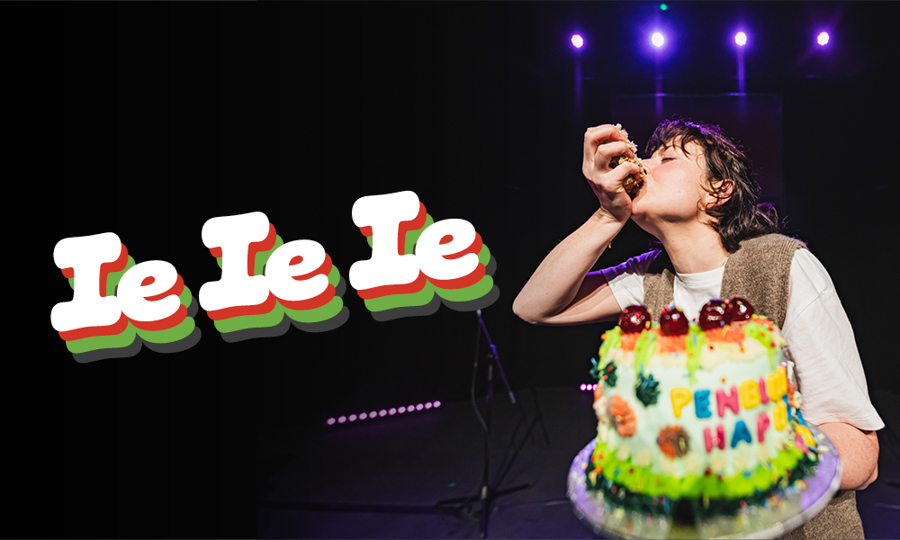 A person holds an extravagant birthday cake with cherries on top and fondant letters saying PENBLWYDD HAPUS. They are holding a handful of cake to their mouth. There are four spotlights above their heads.