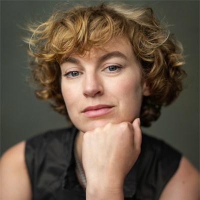 Headshot of Heledd Gwynn. A white woman in her mid-thirties with short mousey coloured curly hair. She is resting her chin on her closed fist, and looking directly into the camera with half a smile on her face.