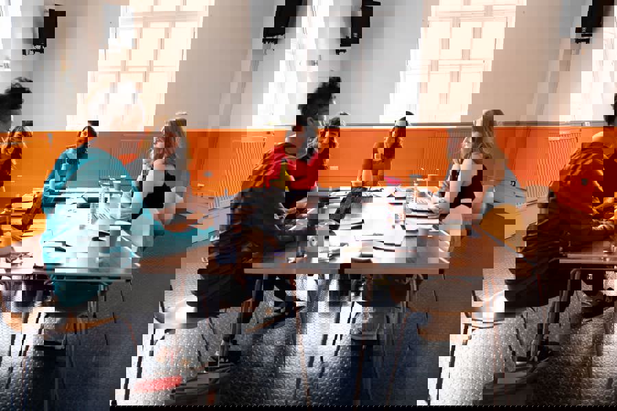 Image of Bren Calon Fi rehearsals. 4 women are sat around a table. Lowri Morgan is a young white woman with blonde hair. Rhiannon Mair is a woman in her thirties with shoulder-length brown hair and wearing and orange jumper. Rhian Blythe is a white woman in her forties with shoulder-length reddy-brown hair. Bethan Marlow is a woman in her thirties with very short brown hair. They are all sitting around a table listening as Bethan speaks. 