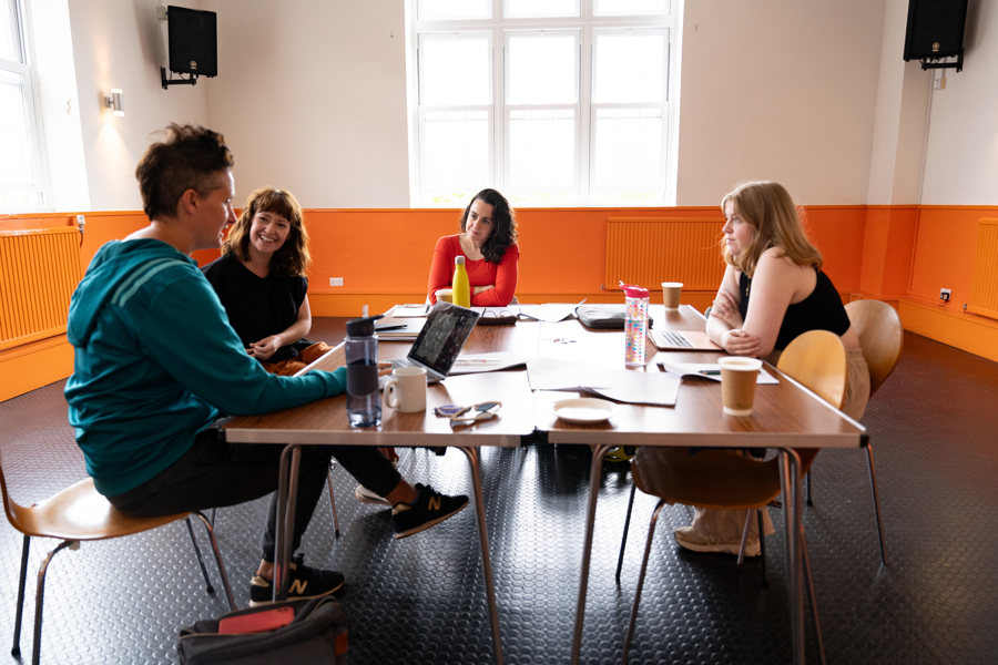 Image of Bren Calon Fi rehearsals. 4 women are sat around a table. Lowri Morgan is a young white woman with blonde hair. Rhiannon Mair is a woman in her thirties with shoulder-length brown hair and wearing and orange jumper. Rhian Blythe is a white woman in her forties with shoulder-length reddy-brown hair. Bethan Marlow is a woman in her thirties with very short brown hair. They are all sitting around a table listening as Bethan speaks. 