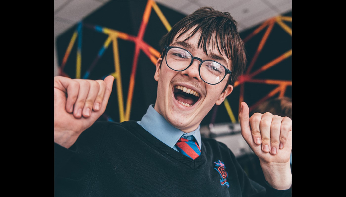 An image of a school boy wearing glasses with brown hair and is smiling with his hands up in the air. 