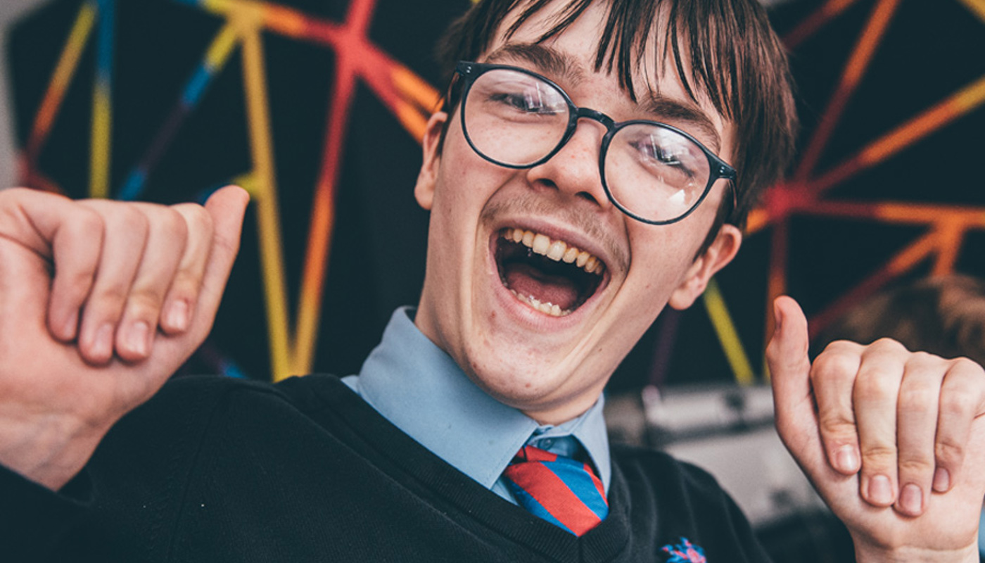 An image of a school boy wearing glasses with brown hair and is smiling with his hands up in the air. 