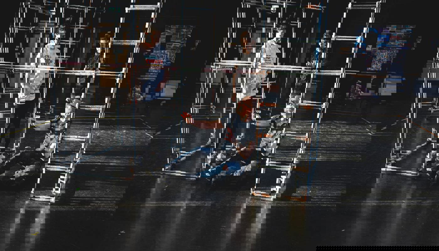 A young woman stands inside a metal structure built from poles. She is not smiling and is looking down at a man. The man is sat with his legs and arms wide on the floor and looks up at the woman. Behind them outside of the structure another woman stands. She looks towards them both.