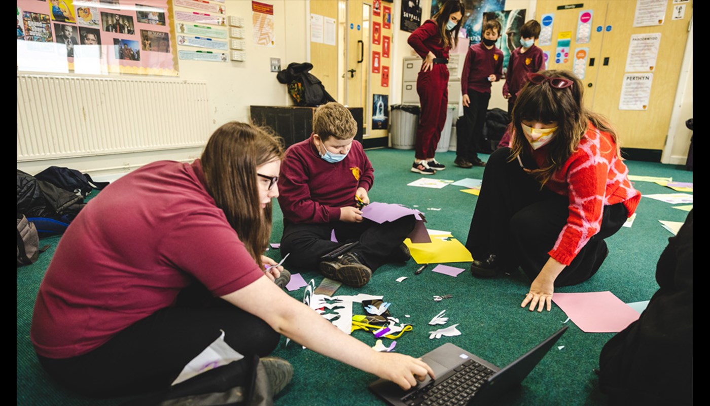 Two young pupils in school uniform and a young woman in formal clothing are sitting on the floor of a classroom. There are pieces of paper and craft supplies on the floor around them. Two are staring down at a piece of work one of the pupils has created, and one is cutting a shape from paper using scissors.