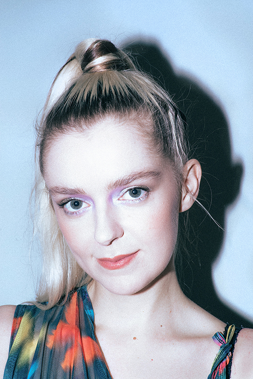 Headshot of Casi Wyn. She is a white woman in her early thirties with blue eyes and blonde hair tied up into a ponytail at the top of her head. She is smiling lightly and looking directly into the camera.