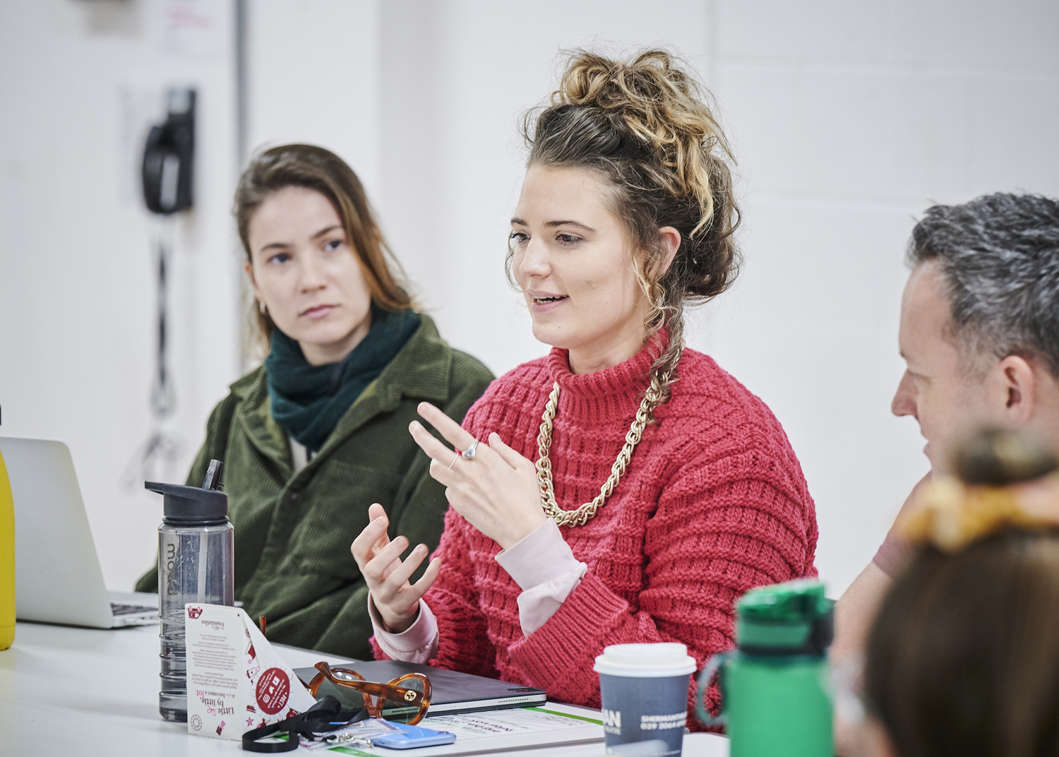 Two young women sit next to each other on a table. One woman is talking and the other is looking at ther. A man is sat beside them also looking at the woman who is talking.