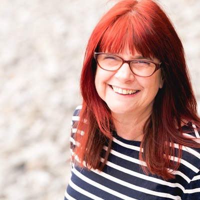 Elanor Higgins's headshot. Elanor is a white female in her forties. She has bright red hair and wears black rimmed glasses. She has on a black and white striped long sleeve shirt. She is looking directly into the camera and smiling with her teeth