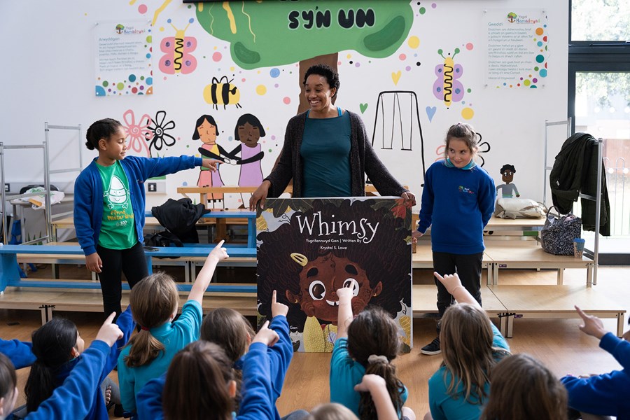 Image of Krystal S Lowe at Swyn workshops. She is a black woman in her thirties with afro hair. She wears a turquoise tshirt with a brown cardigan. She is holds a large copy of the book Whimsy, with an illustration of a young black girl. On the left, there is a mixed race young girl with short afro hair tied into small buns at the back of her head. She wears a green Mr Urdd tshirt with a blue cardigan on top. The young girl on the other side is white with brown hair tied into a french plait on her head.