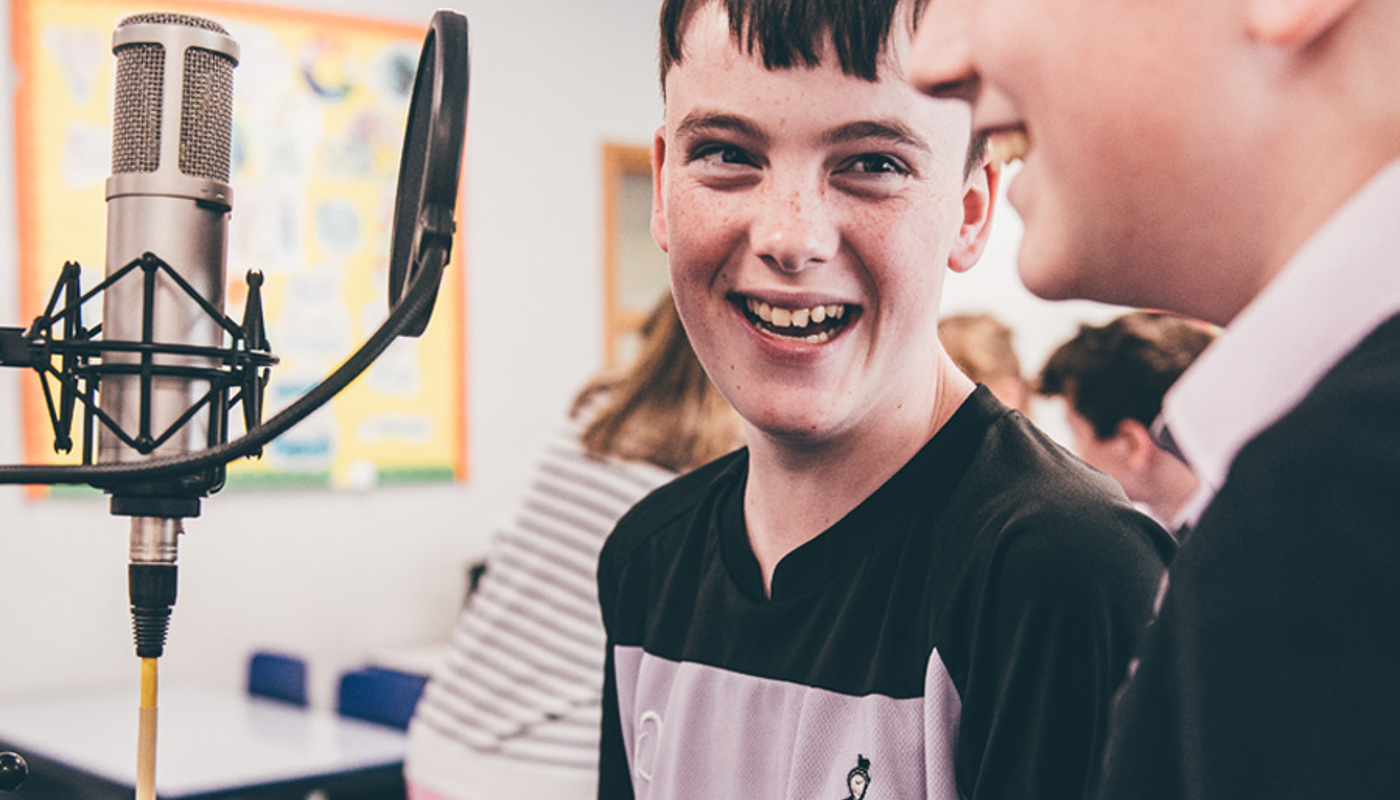 An image of two boys laughing and smiling at each other behind a microphone. They are both wearing dark navy clothing. 