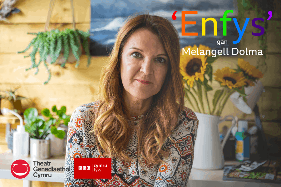 Woman sits in front of a wooden wall. She sits in the centre of the frame and is staring directly into the camera. Behind her there is a white table covered in various small plants in pots and a jug of yellow sunflowers. Multi-coloured text on the image reads translated ‘Rainbow by Melangell Dolma’