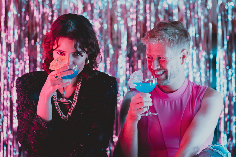 Poster image for Byth Bythoedd Amen. Two people are sitting in front of a foil curtain drinking blue cocktails - a woman wearing all black is drinking from the glass, while a man wearing pink grins at her.