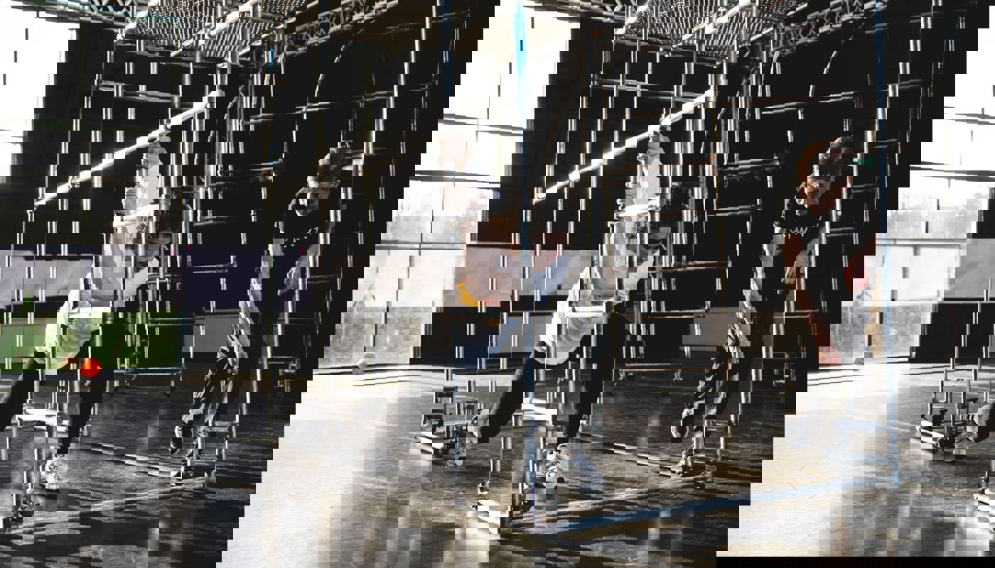 Young woman stands inside a metal structure built from long poles. She is lunging forwards with her hands above her head grabbing on to one of the poles. Another woman stands to the side, she is crouched down and looking towards the young woman.