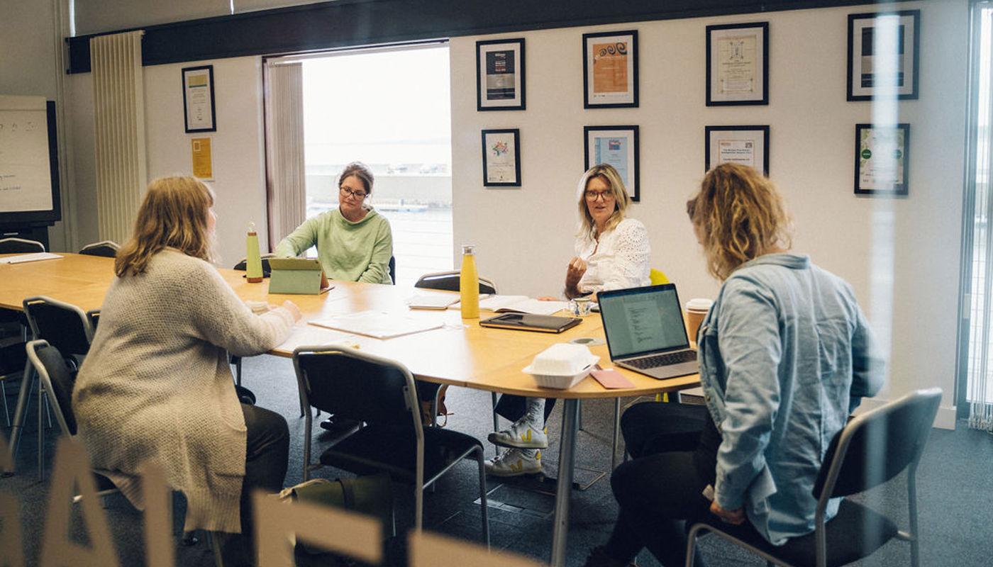 5.	4 women sit at a table. Two women have their backs to the camera. The two other women face the camera. One of them is talking to another and has her mouth half-open. The other woman is writing something on a piece of paper. There is a takeaway box, water bottles, notebooks, a laptop and pens on the table.