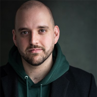 Headshot of Mark Henry Davies. He is a white man. He is bald and has short stubble around his chin and mouthg. He is wearing a dark green hoodie with a black jacket over it. 