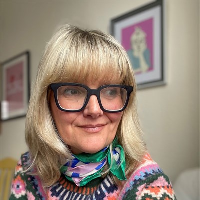 A headshot of Iola Ynyr. She is a white middle aged woman with shoulder-length blonde hair. She wears large, square black rimmed glasses. She is looking off to the side and has a slight smile on her face. She is wearing a colourful fair isle