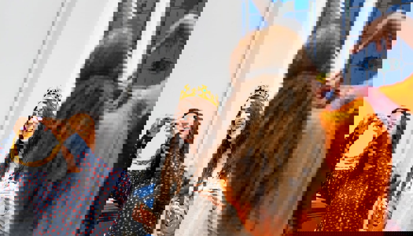 Three people in a room, one woman has her back to the camera. One of the women facing the camera is smiling and is wearing a plastic crown on her head. The other woman facing the camera is putting an accessory in her hair and is also smiling.