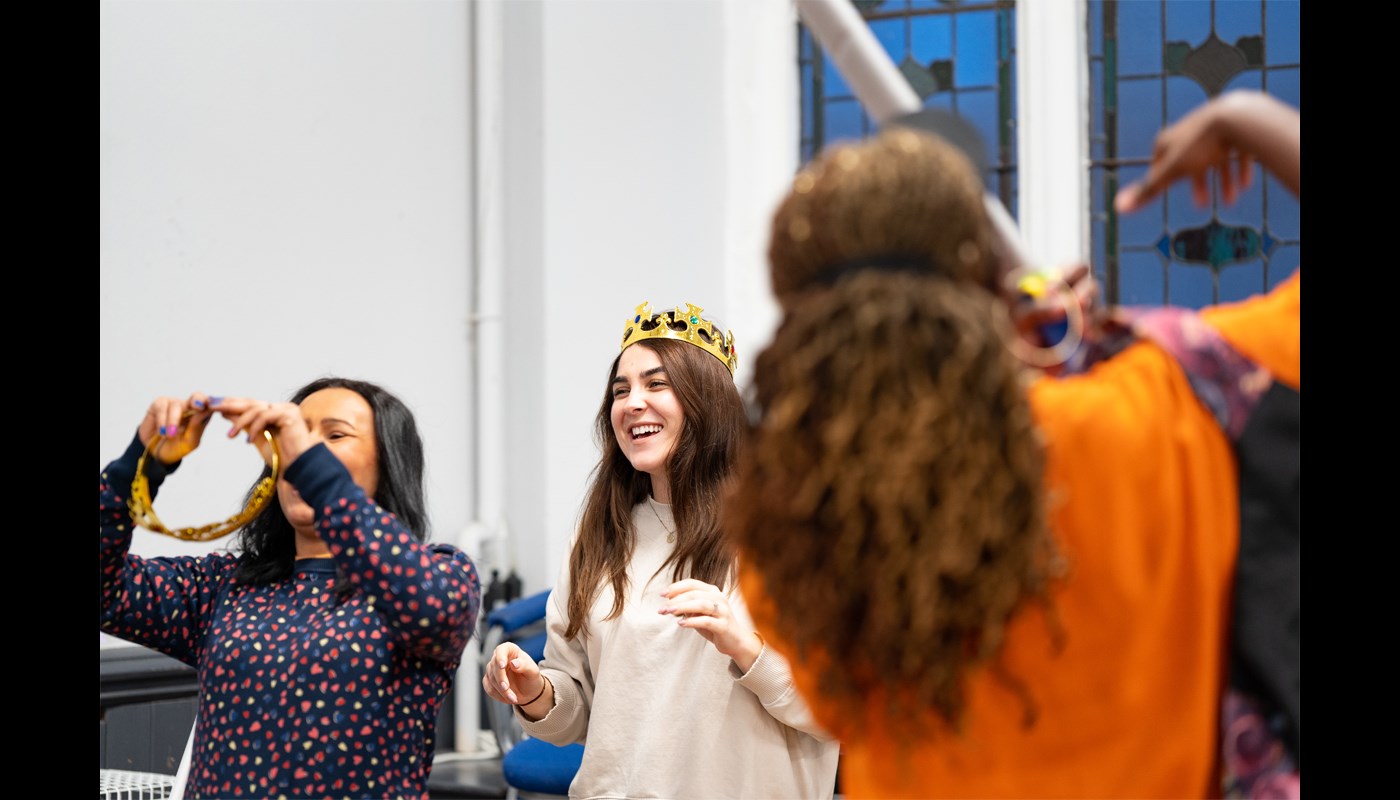 Three people in a room, one woman has her back to the camera. One of the women facing the camera is smiling and is wearing a plastic crown on her head. The other woman facing the camera is putting an accessory in her hair and is also smiling.