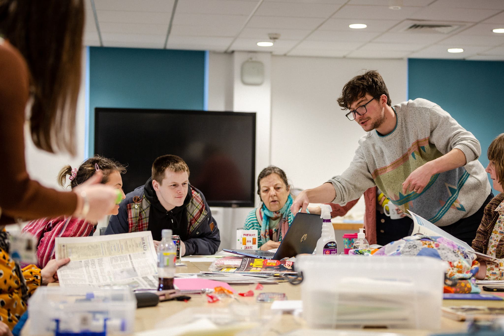 4 people sat round a table. On the table different arts and crafts materials and scraps of paper are scattered. One of the people is standing up looking down at another person's work and pointing at it. 