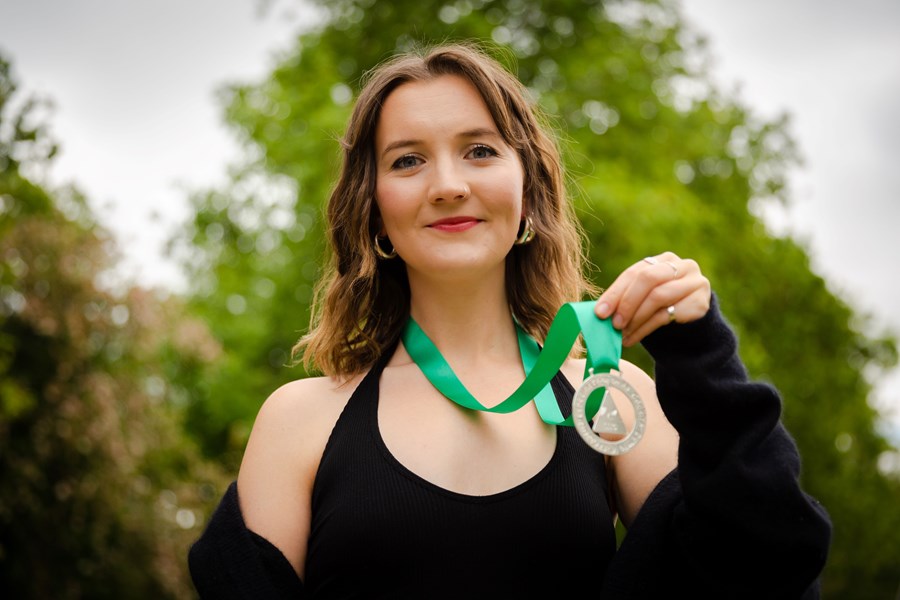 An image of a woman wearing an Urdd medal around her neck and is smiling into the camera. She is wearing a black top and black cardigan. The image is taken outside.
