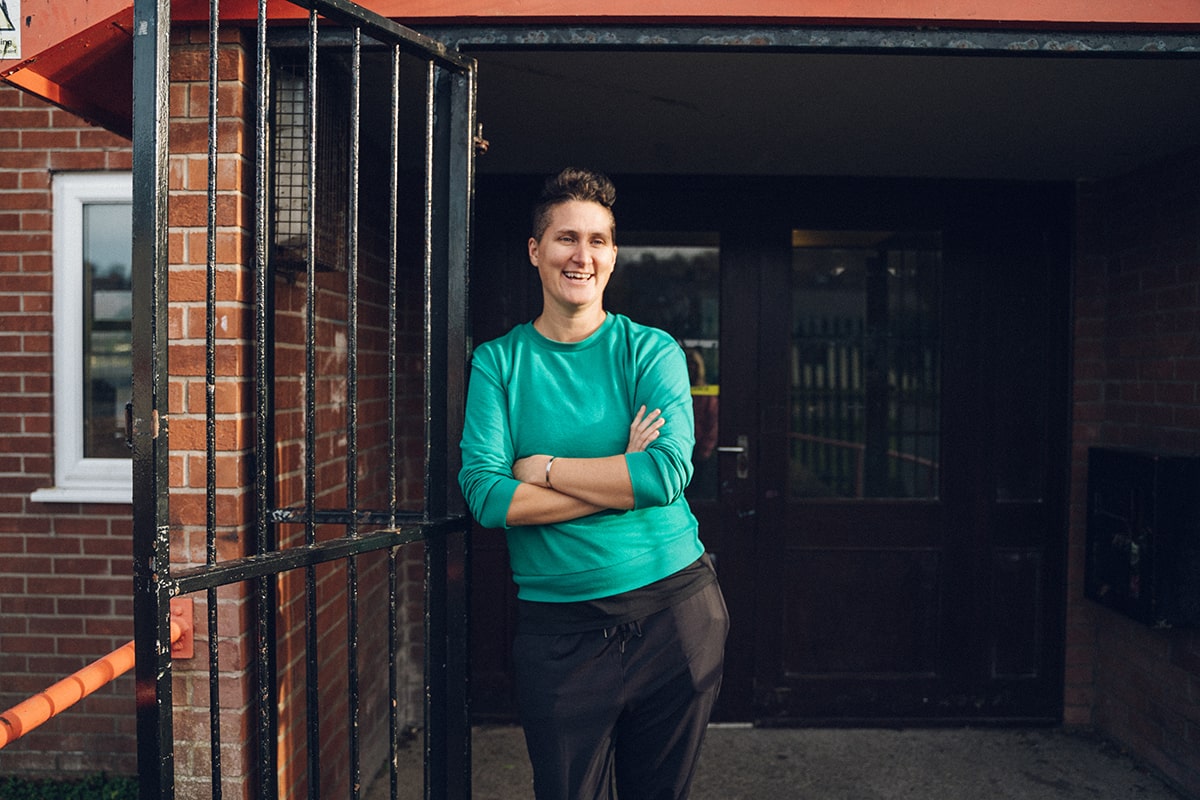 Woman leans against a black iron gate in the doorway to a building. She is smiling and is looking off camera.
