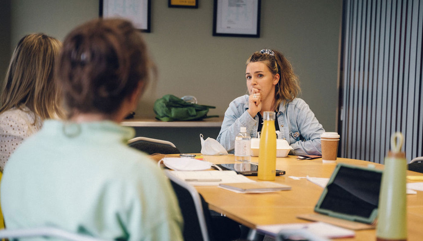 3.	Young woman is sat at a table facing the camera. Two other women sit in front of her with their backs to the camera. Her eyebrows are raised and she covers her mouth with her hand. There are water bottles, coffee cups, pieces of paper and pens on the table. 