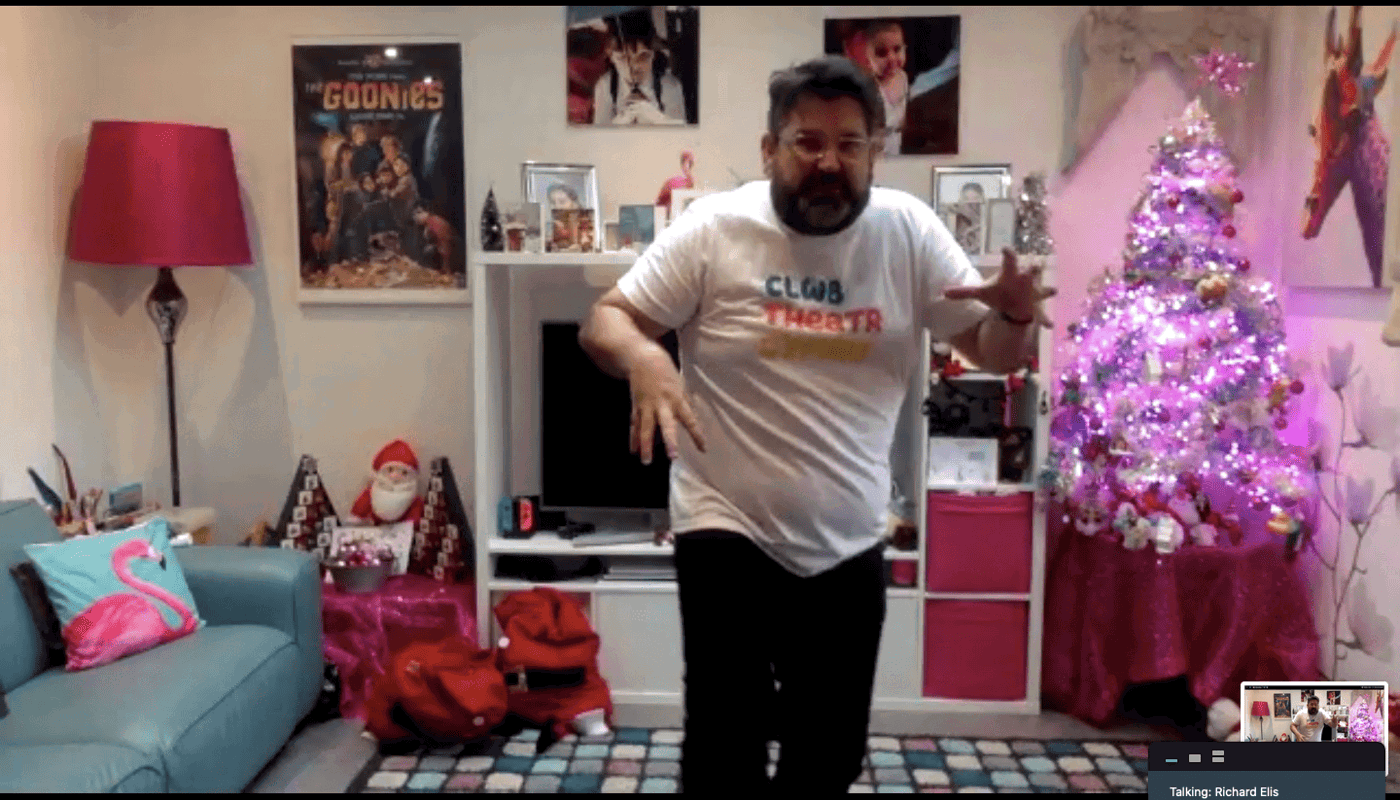 1.	Screenshot of a Zoom recording. Man is stood in a room. He is staring at the camera. His back is hunched over one of his hands is lowered and the other is raised. He is looking at the camera. Behind him there is a sofa, lamp, movies posters, shelves and other various images. There is also a table with two advent calendars and a Santa Clause teddy on, as well as another table with a Christmas tree decorated with baubles and fairy lights.