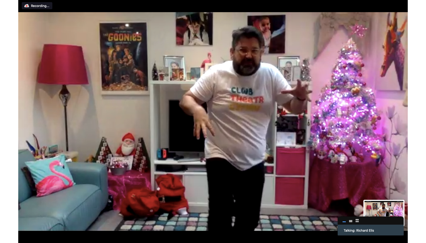 1.	Screenshot of a Zoom recording. Man is stood in a room. He is staring at the camera. His back is hunched over one of his hands is lowered and the other is raised. He is looking at the camera. Behind him there is a sofa, lamp, movies posters, shelves and other various images. There is also a table with two advent calendars and a Santa Clause teddy on, as well as another table with a Christmas tree decorated with baubles and fairy lights. 