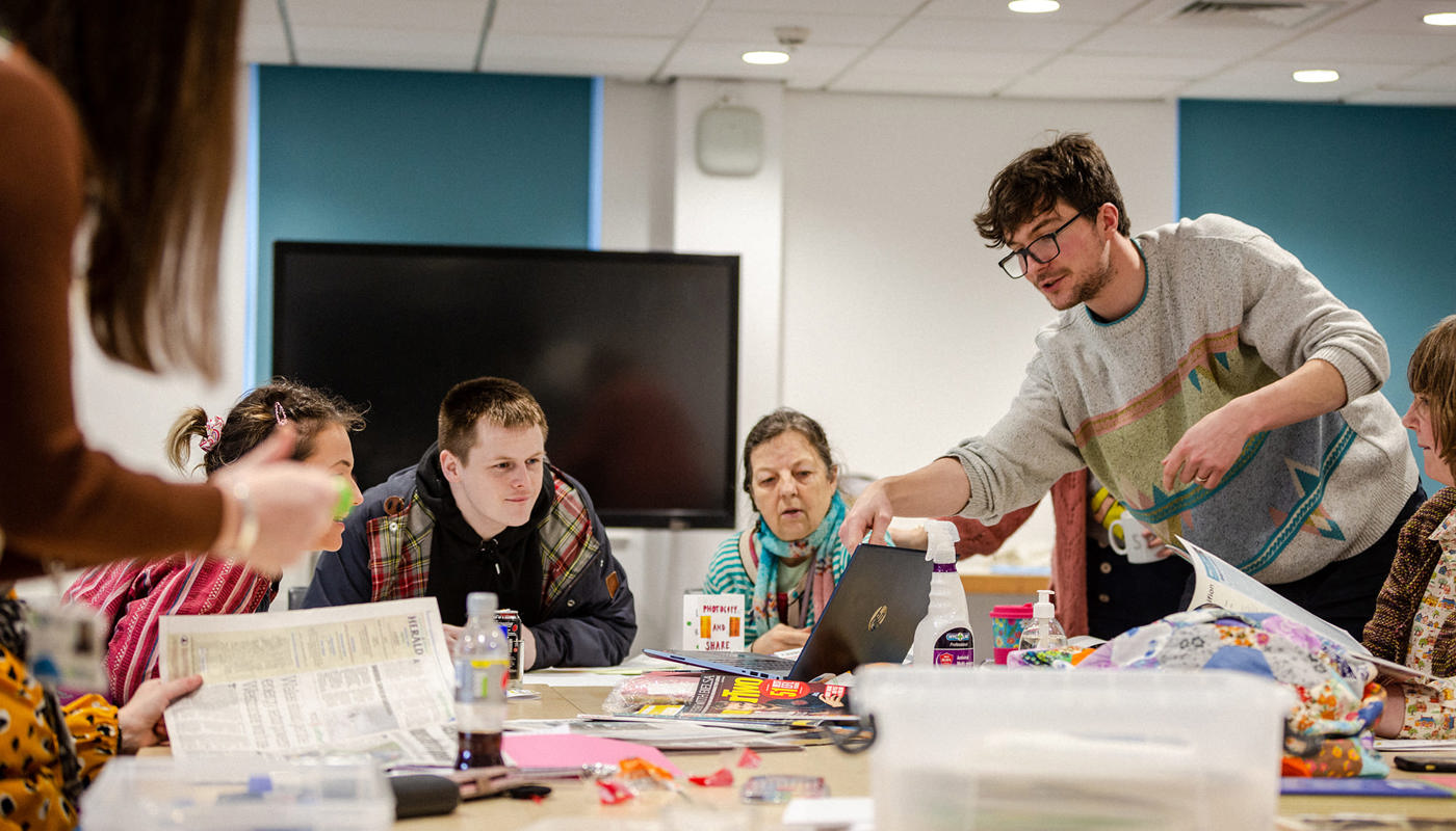 4 people are sat down at a table. There is a man stood leaning over them pointing towards a laptop screen in front of them. They are all looking towards the laptop screen. On the table there are various materials scattered and pieces of colourful paper cut up.