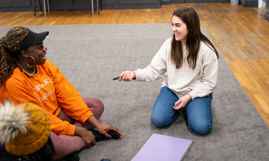 Two women sat on the floor. Both are smiling and looking towards each other. There is a purple piece of paper between them and one is holding a pen in her hand. 